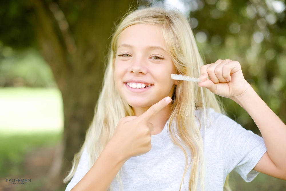 Smiling young girl pointing at her teeth outdoors—early orthodontic consultation helps determine Invisalign cost in New Braunfels, TX.