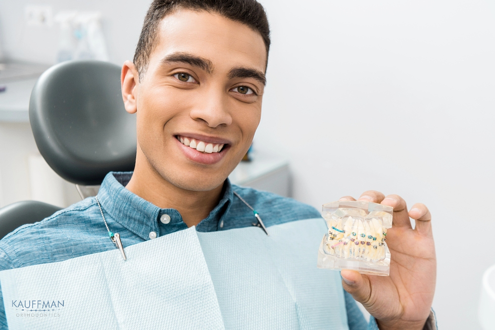 Smiling man holding a dental model with braces while sitting in a dental chair, showcasing orthodontic treatment options after understanding about How Long Do Braces Take in New Braunfels, TX